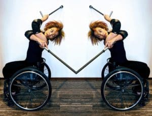 Alice Sheppard seated in her wheelchair with two crutches, crosses her body and arches her spine to look behind. The photo is a mirror reflection and Alice looks back at herself as her crutches disappear into each other in a seamless touchpoint. Photo by Lisa Niedermeyer. Caption Text: Alice Sheppard from solo work Where Good Souls Fear. Photo by Lisa Niedermeyer.