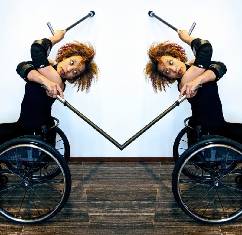 Alice Sheppard seated in her wheelchair with two crutches, crosses her body and arches her spine to look behind. The photo is a mirror reflection and Alice looks back at herself as her crutches disappear into each other in a seamless touchpoint. Photo by Lisa Niedermeyer. Caption Text: Alice Sheppard from solo work Where Good Souls Fear. Photo by Lisa Niedermeyer.