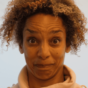Alice Sheppard, a light skinned multiracial Black woman with brown eyes and blonde, mustard, and brown striped curly hair, wears a peach colored sweatshirt. In this video, she performs a face dance moving in and out of a series of intense facial expressions to create a dance capturing some of the feelings of forgetting. In between some of these expressions, an AI intervenes. Time slows as we watch the AI create the transition, by changing Alice’s facial features and interpreting her face through its own lens. In this still her eyebrows are lifted and she looks both uncomfortable and surprised.